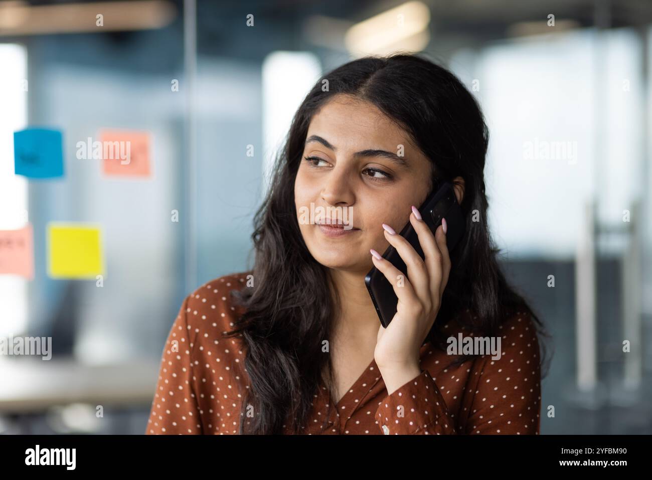 Junge hispanische Geschäftsfrau in moderner Büroumgebung am Telefon, die Fokus ausdrückt. Professionelle Kleidung und Haftnotizen im Hintergrund deuten auf eine kreative Atmosphäre am Arbeitsplatz hin. Stockfoto