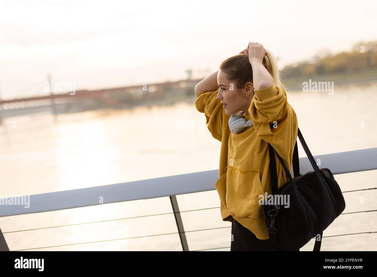 Bei Sonnenuntergang steht eine fröhliche junge Frau am Wasser und genießt einen ruhigen Moment, während sie spielerisch ihre Haare zurückzieht und fühlt Stockfoto