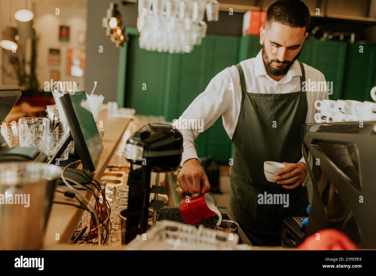 In einem warmen und einladenden Café gießt ein engagierter Barista fachmännisch Milch in eine Kaffeetasse und sorgt so für ein angenehmes Erlebnis für Gäste. Stockfoto
