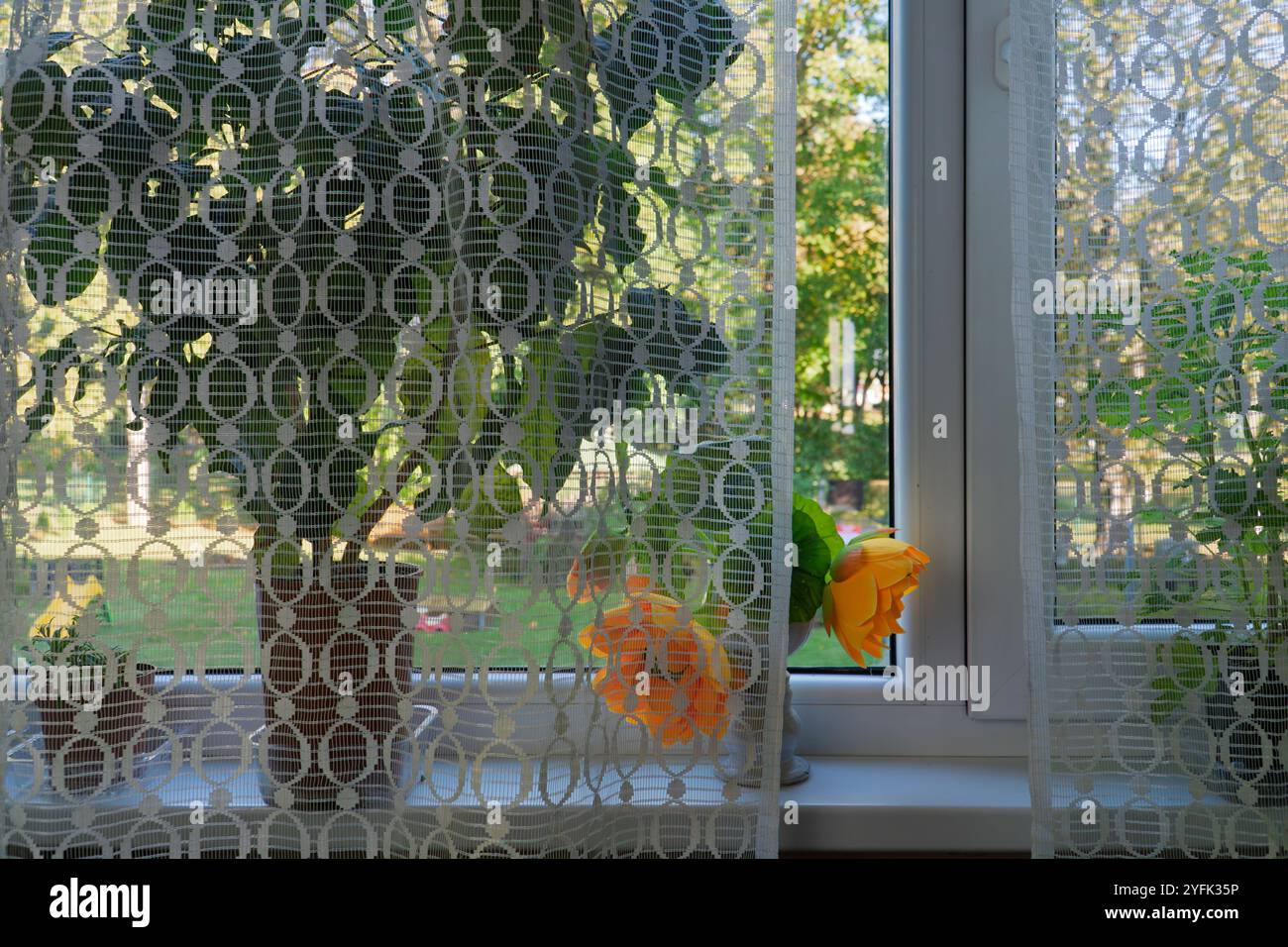 Diese bezaubernde Fensterbank zeigt lebensechte künstliche Blumen, die dem Raum einen Farbakzent verleihen. Die zarten Spitzenvorhänge und Topfpflanzen Stockfoto