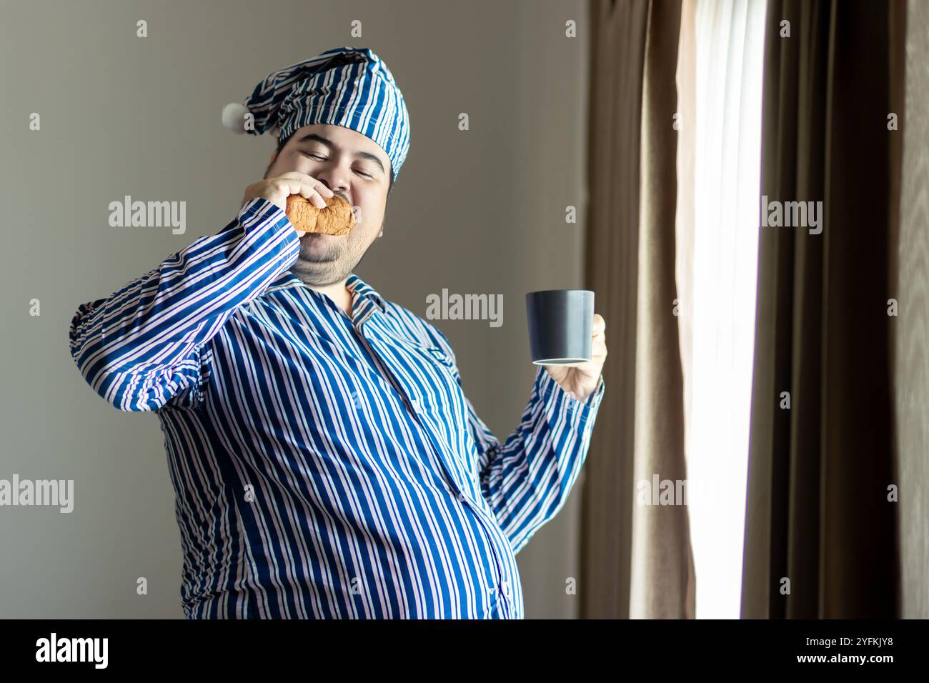 Fette Mann im Pyjama, die am Fenster frühstücken Stockfoto