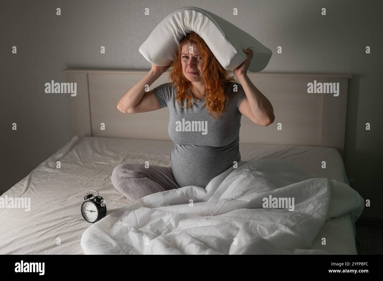 Eine kaukasische Schwangere sitzt im Bett, hält ein Kissen auf dem Kopf und schreit. Schlaflosigkeit. Stockfoto