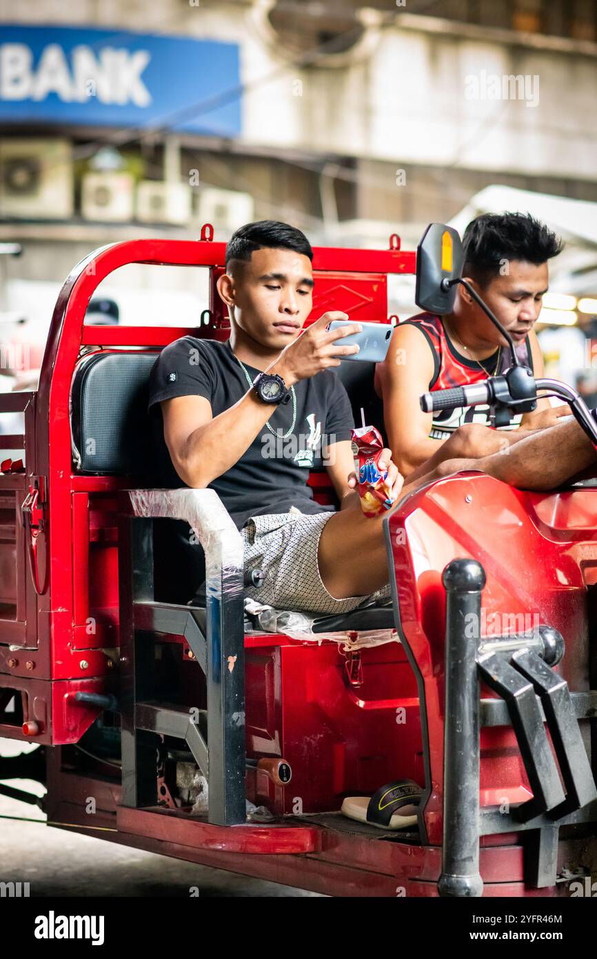 Zwei junge philippinische Arbeiter legen eine Pause ein und saßen auf ihrem Jeep oder Trike auf einem geschäftigen Markt in Manila, den Philippinen. Stockfoto