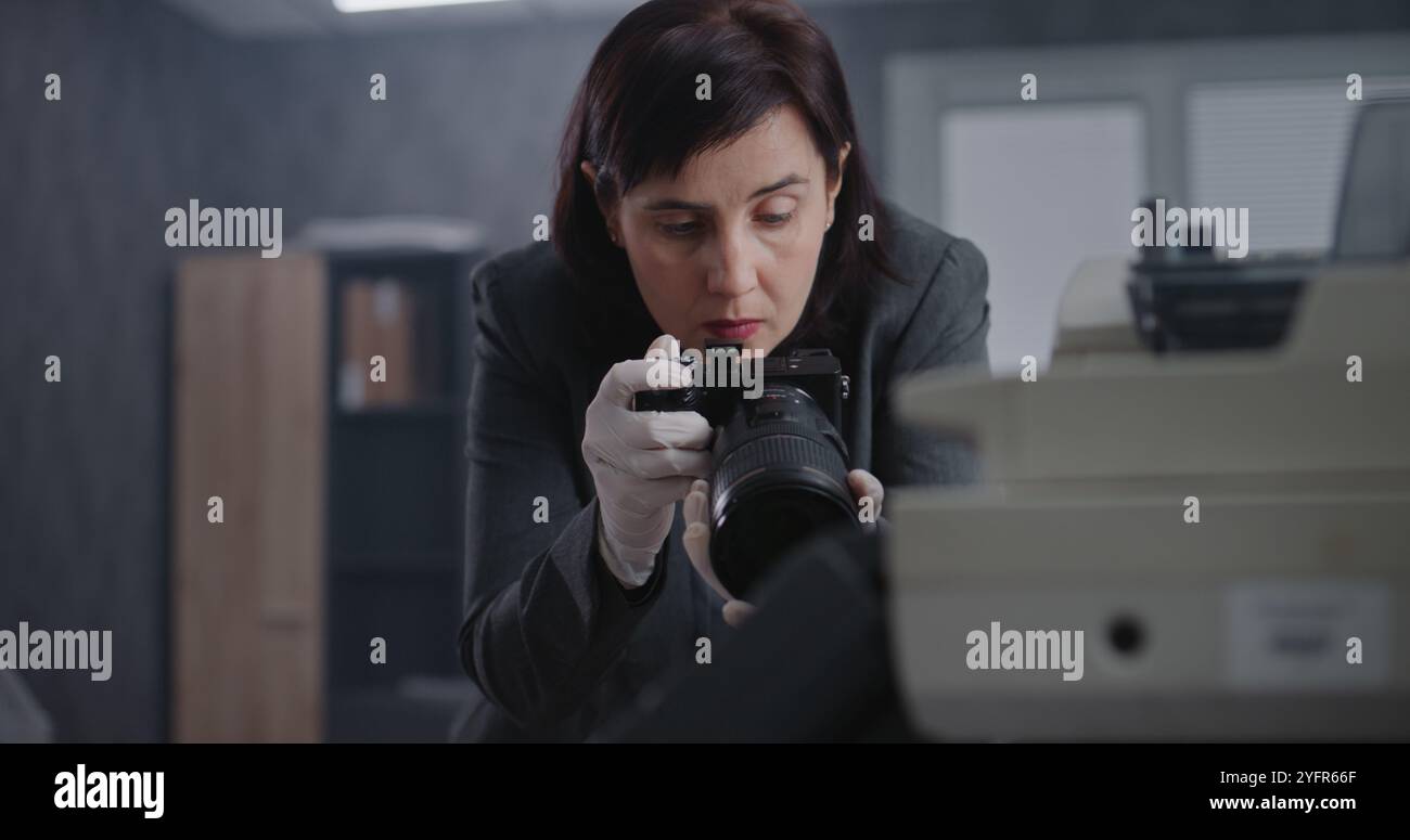 Büroraub oder Mordfall: Eine Kriminalistin fotografiert Beweise am Tatort mit Kamera. Afroamerikanischer Polizist und Detective im Hintergrund. Das Untersuchungsteam arbeitet. Stockfoto