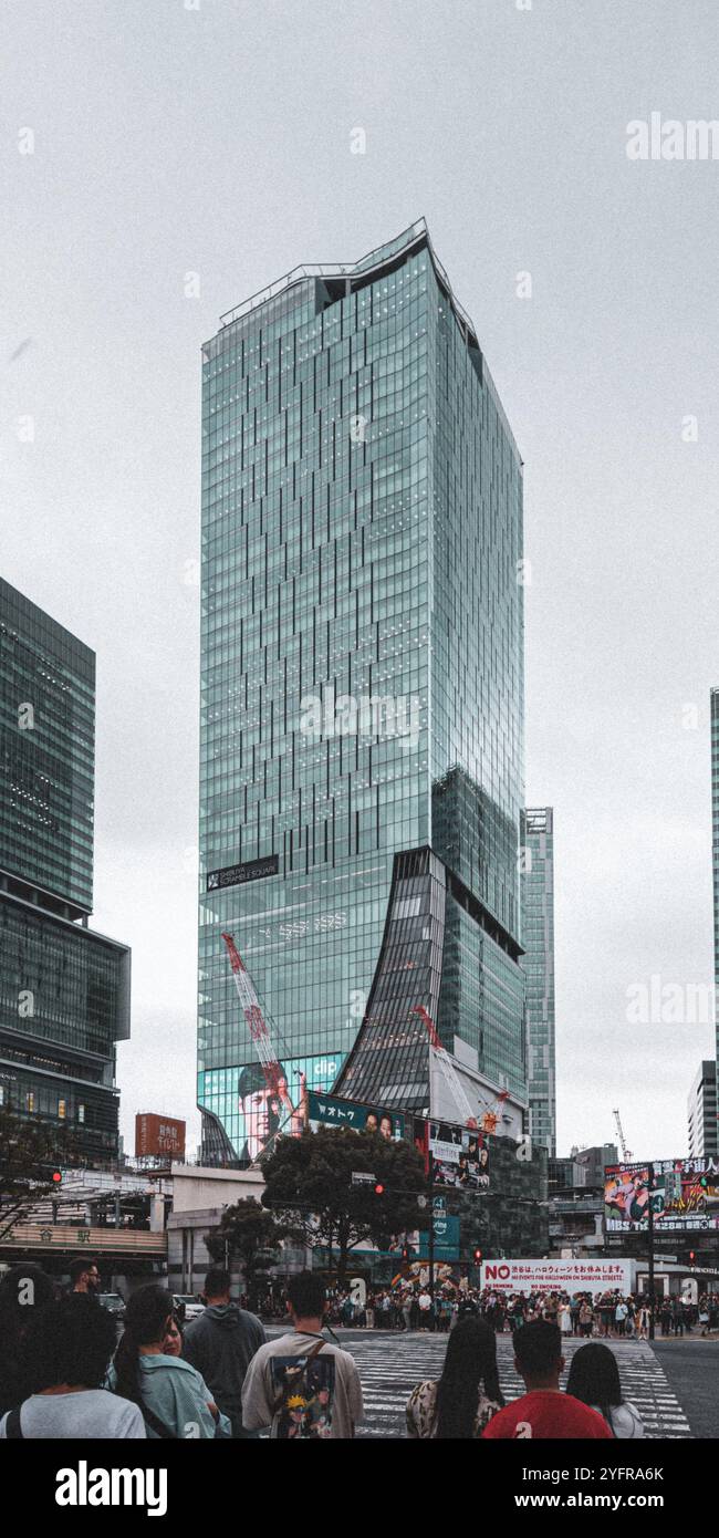 Moderner Glashochhaus in einem belebten Stadtgebiet, umgeben von Geschäften und Fußgängern an einem bewölkten Tag Stockfoto