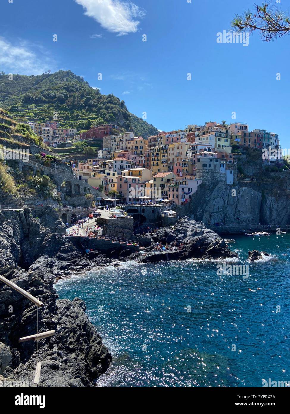 Seitlicher Blick auf die Stadt Manarola, Cinque Terre, Ligurien, Italien, mit ihrem blauen Meer und farbenfrohen Gebäuden und umliegenden Hügeln, an einem sonnigen Sommertag - Smartphone-aufgenommenes Stockfoto