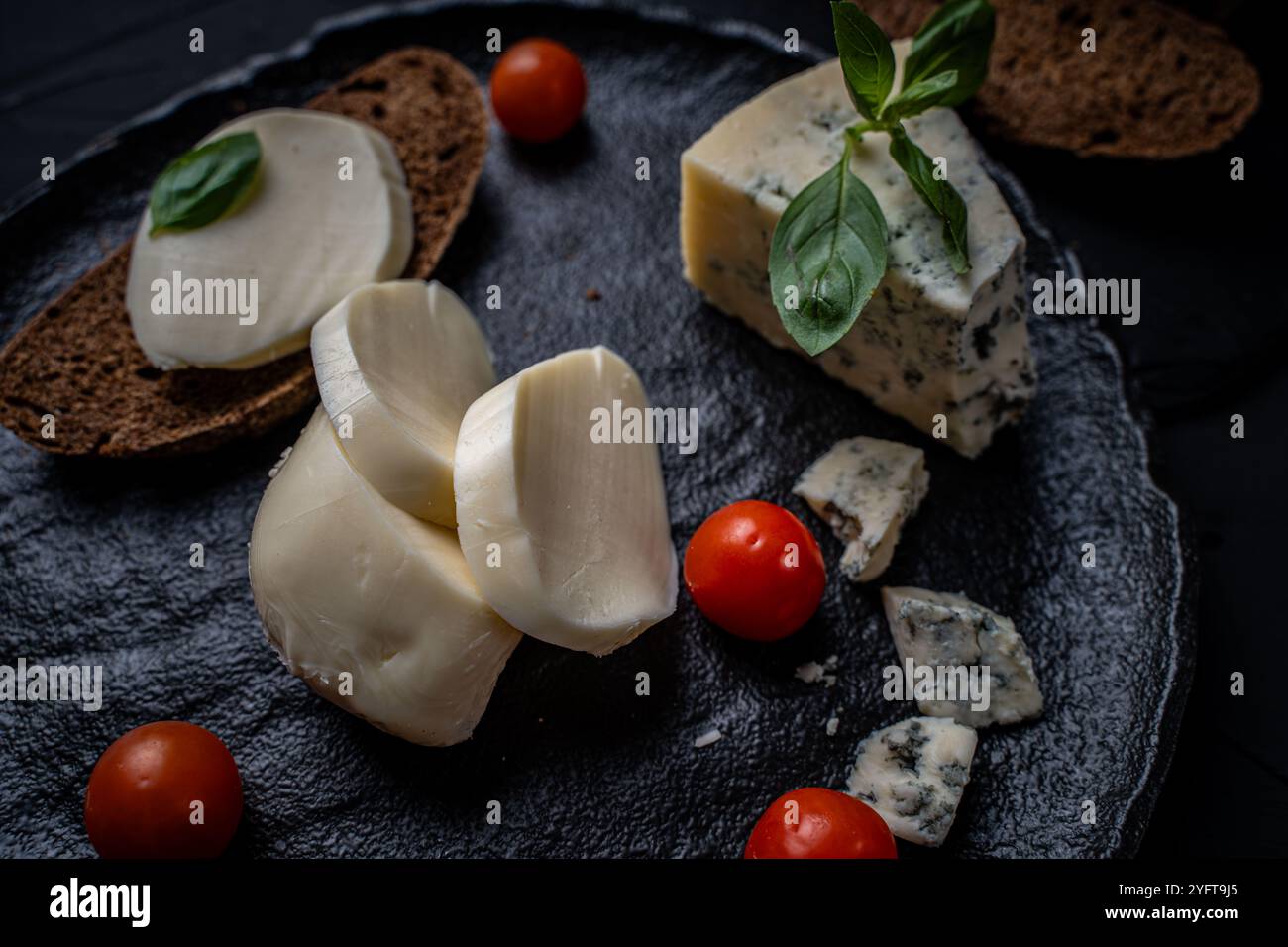 Mozzarella und blaue Garganzola mit dunklem Roggenbrot und Kirschtomaten mit grünem Basilikum auf schwarzer Glimmerplatte auf dunklem Hintergrund. Hohe Qualität Stockfoto