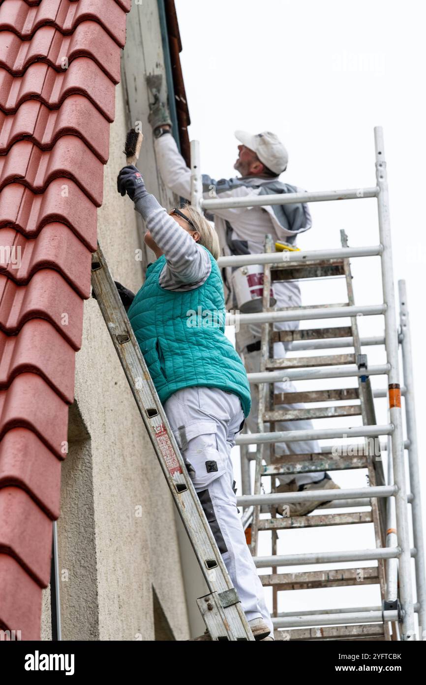 Malerin kratzt mit einer Drahtbürste die alte Farbe auf denHolzteilen, dem Dachüberstand an einem Haus ab. Sie stehen auf einer Leiter, Vorarbeiten zum Malern des Dachüberstands, Modell freigegeben für redaktionelle Nutzung, Dachüberstand streichen *** Malerin kratzt mit einem Drahtpinsel die alte Farbe auf den Holzteilen ab, den Dachüberhang auf einem Haus Sie steht auf einer Leiter, Vorarbeiten zum Bemalen des Dachüberhangs, Modell freigegeben für redaktionelle Verwendung, Bemalen des Dachüberhangs 20241105-DSC 8793 Stockfoto