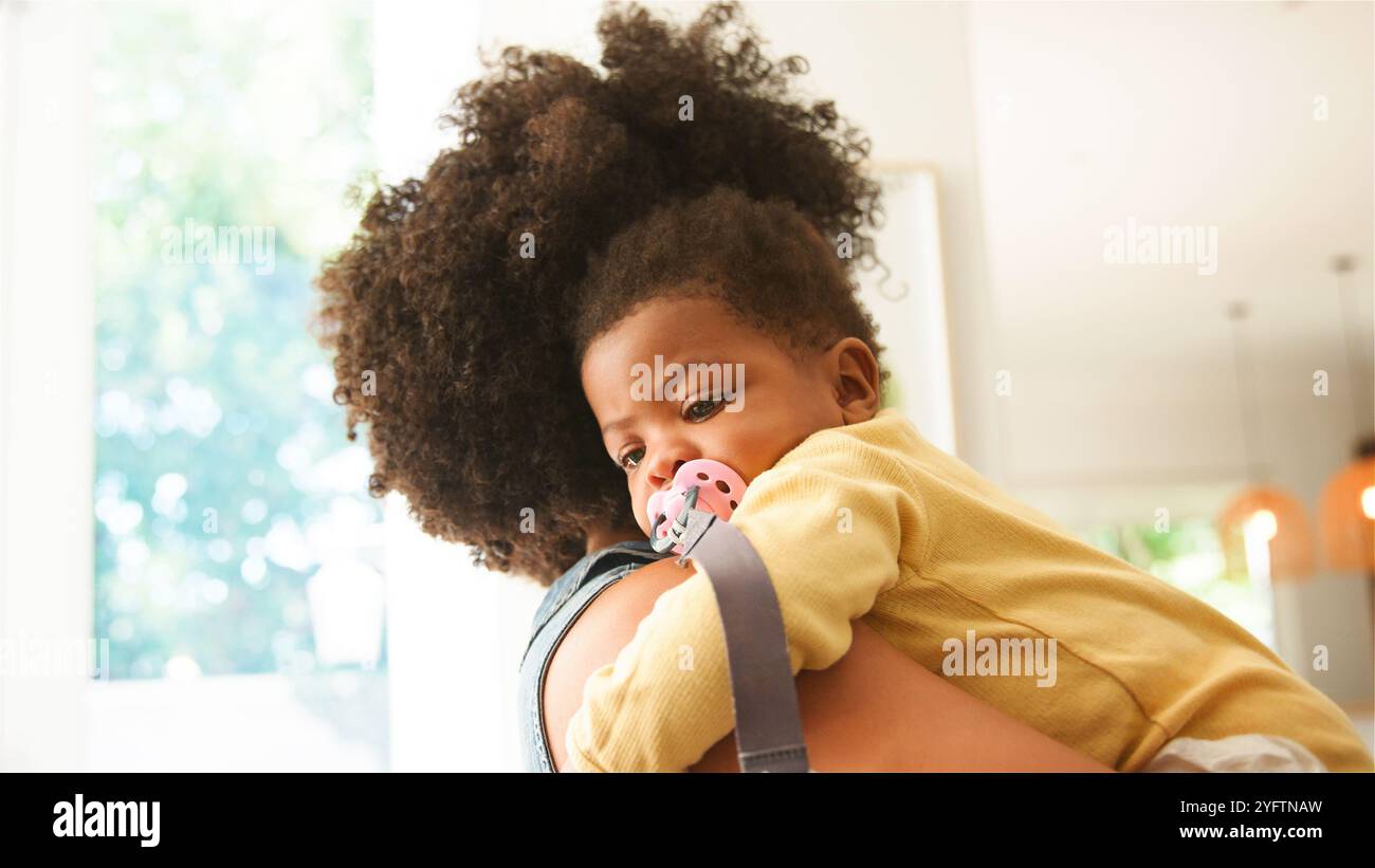 Eine Mutter wiegt ihr Baby in den Armen und bietet Komfort und Sicherheit in einer warmen, einladenden Innenumgebung. Das Baby, mit einem Schnuller, sieht besonnen aus Stockfoto