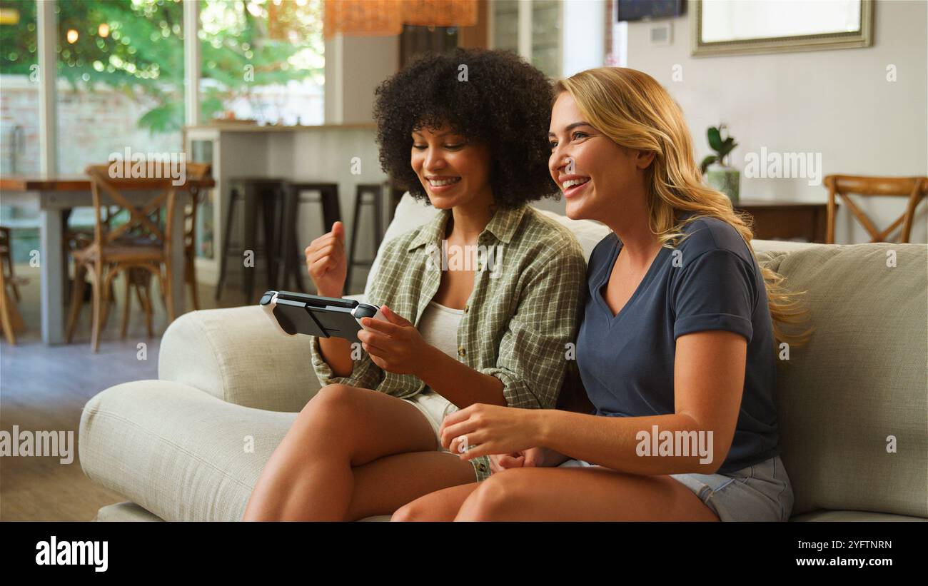 Zwei Freunde sitzen bequem auf einem Sofa in einem hellen Wohnzimmer und teilen sich Lachen und Aufregung, während sie gemeinsam ein Videospiel auf einer Handhel spielen Stockfoto