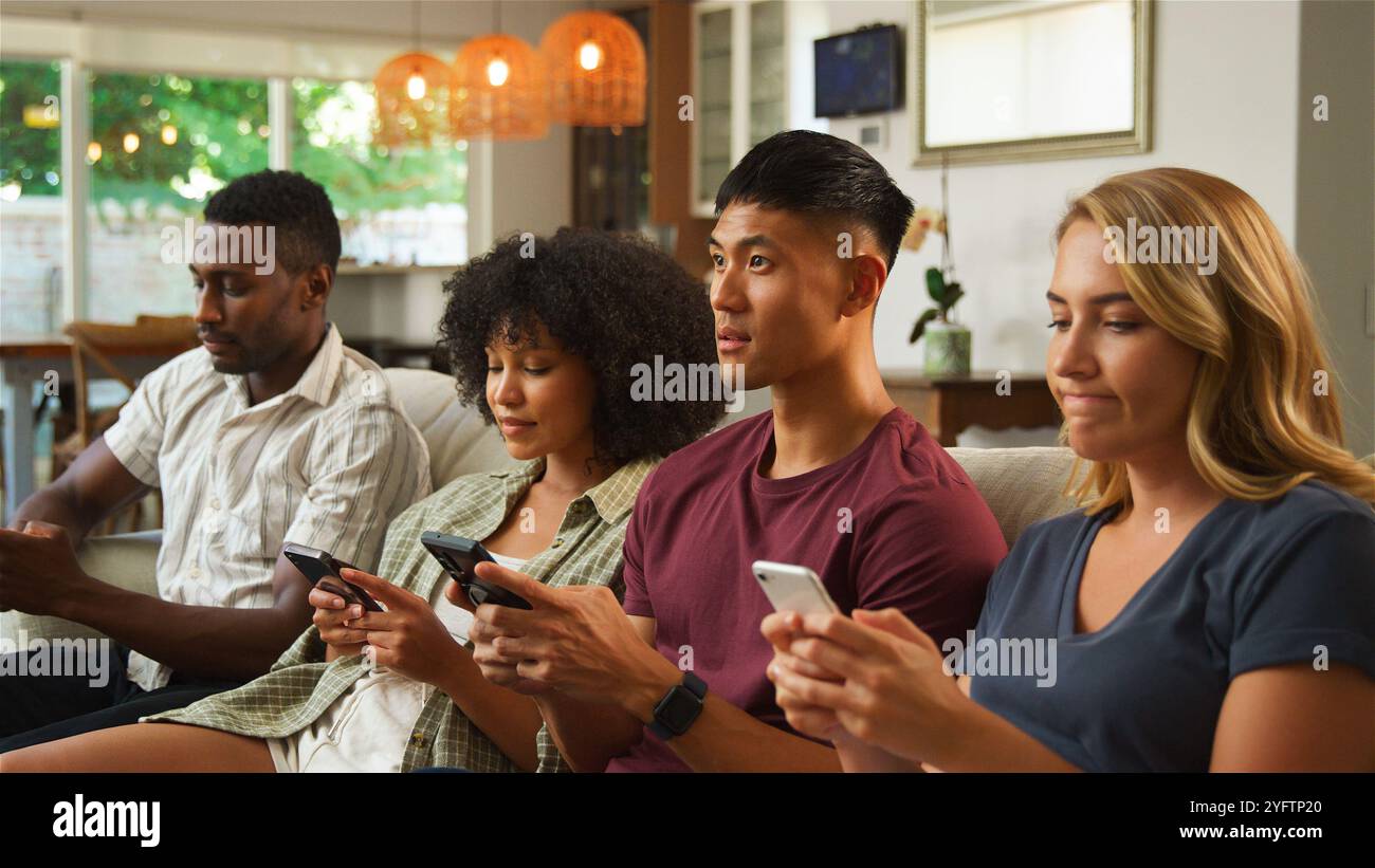 Eine bunte Gruppe von vier Freunden sitzt zusammen auf einer Couch in einem hellen, einladenden Wohnzimmer. Jede Person konzentriert sich auf ihr Smartphone, scheint es Stockfoto