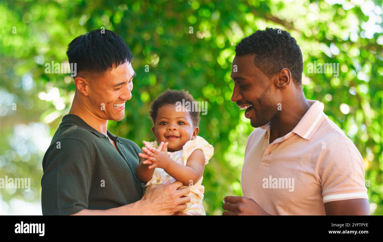 Zwei Väter interagieren fröhlich mit ihrem fröhlichen Baby draußen. Stockfoto