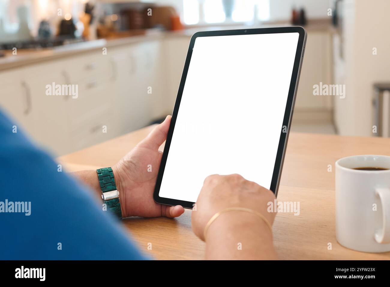 Eine Person verwendet ein Tablet an einem Küchentisch, umgeben von einer gemütlichen Atmosphäre. Sanftes Morgenlicht strömt durch ein Fenster und verbessert das Licht Stockfoto
