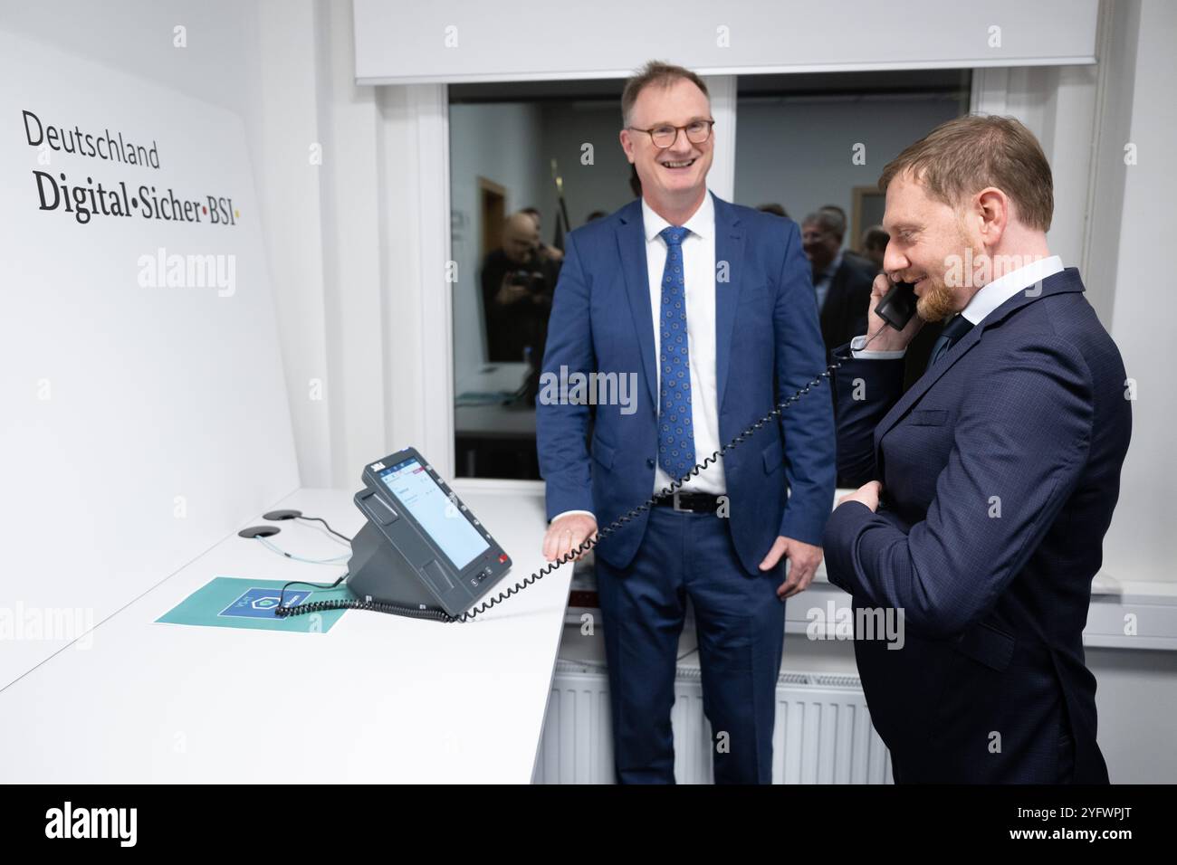 Freital, Deutschland. November 2024. Günther Welsch (l), Leiter der Abteilung Kryptotechnologie, erklärt dem sächsischen Ministerpräsidenten Michael Kretschmer (CDU) ein Telefon für die Hochsicherheits-Telefonielösung zur Übermittlung von Verschlusssachen beim Bundesamt für Sicherheit in der Informationstechnik (BSI). Die Telefonielösung ist für den Testbetrieb in Sachsen vorgesehen. Quelle: Sebastian Kahnert/dpa/Alamy Live News Stockfoto