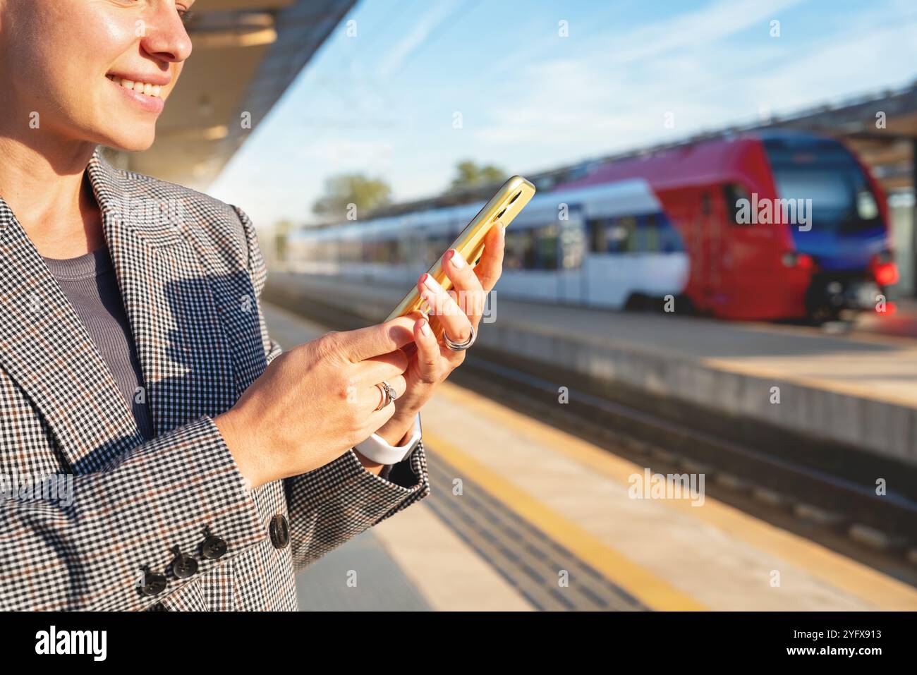 Online-Buchung von Zugtickets. Eine Pendlerin steht auf einem Bahnsteig und benutzt ihr Smartphone mit einem unscharfen Zug im Hintergrund. Stockfoto