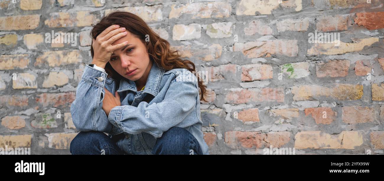 Die überforderte und deprimierte Stadtfrau sitzt draußen in der Stadt neben einer Ziegelmauer mit der Hand auf der Stirn. Stress und psychische Erkrankungen. Stockfoto