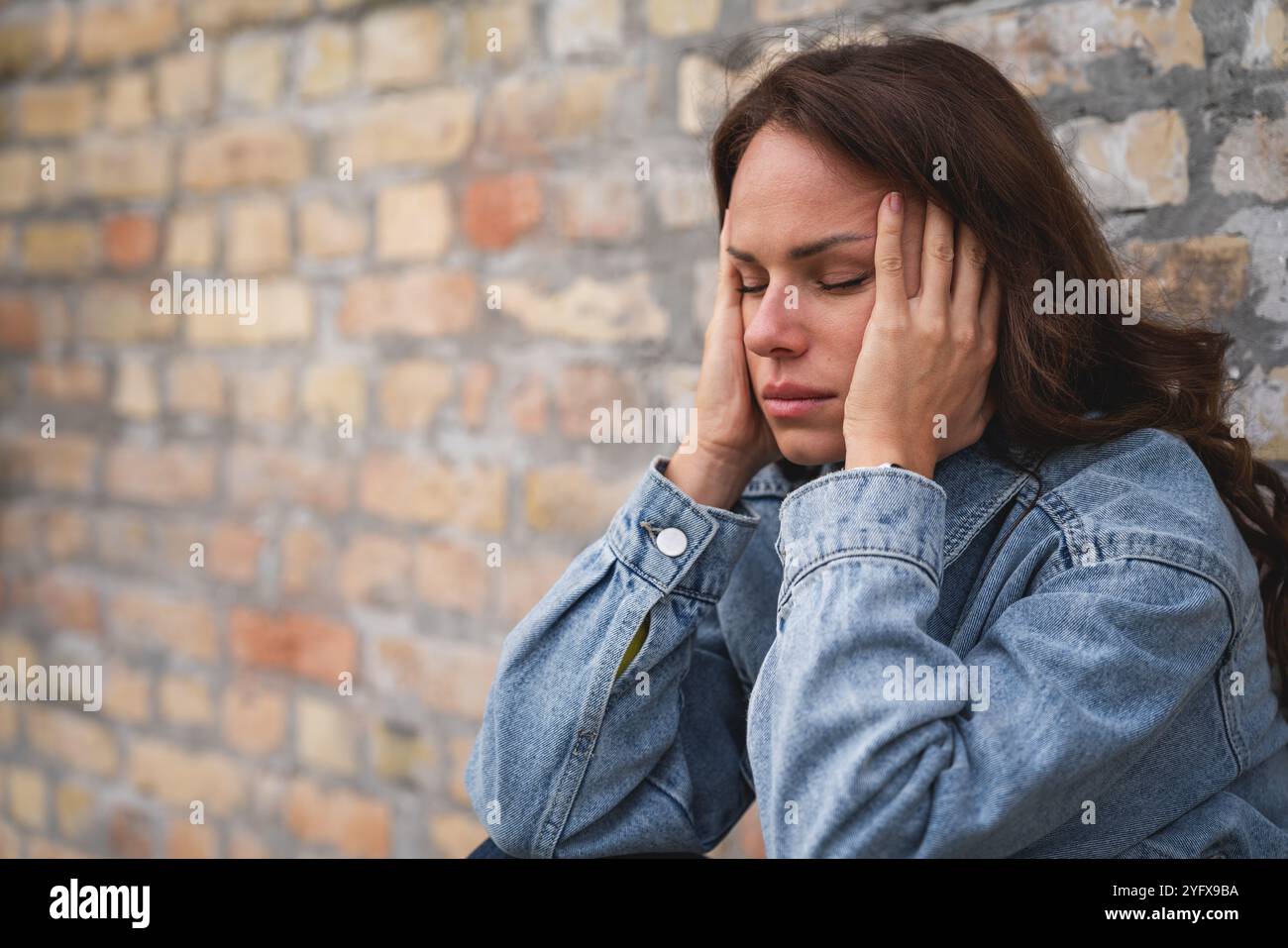 Eine überforderte Frau mit geschlossenen Augen und Kopf in Händen sitzt in der Nähe einer Ziegelmauer auf einer Stadtstraße und vermittelt emotionalen Stress. Stockfoto
