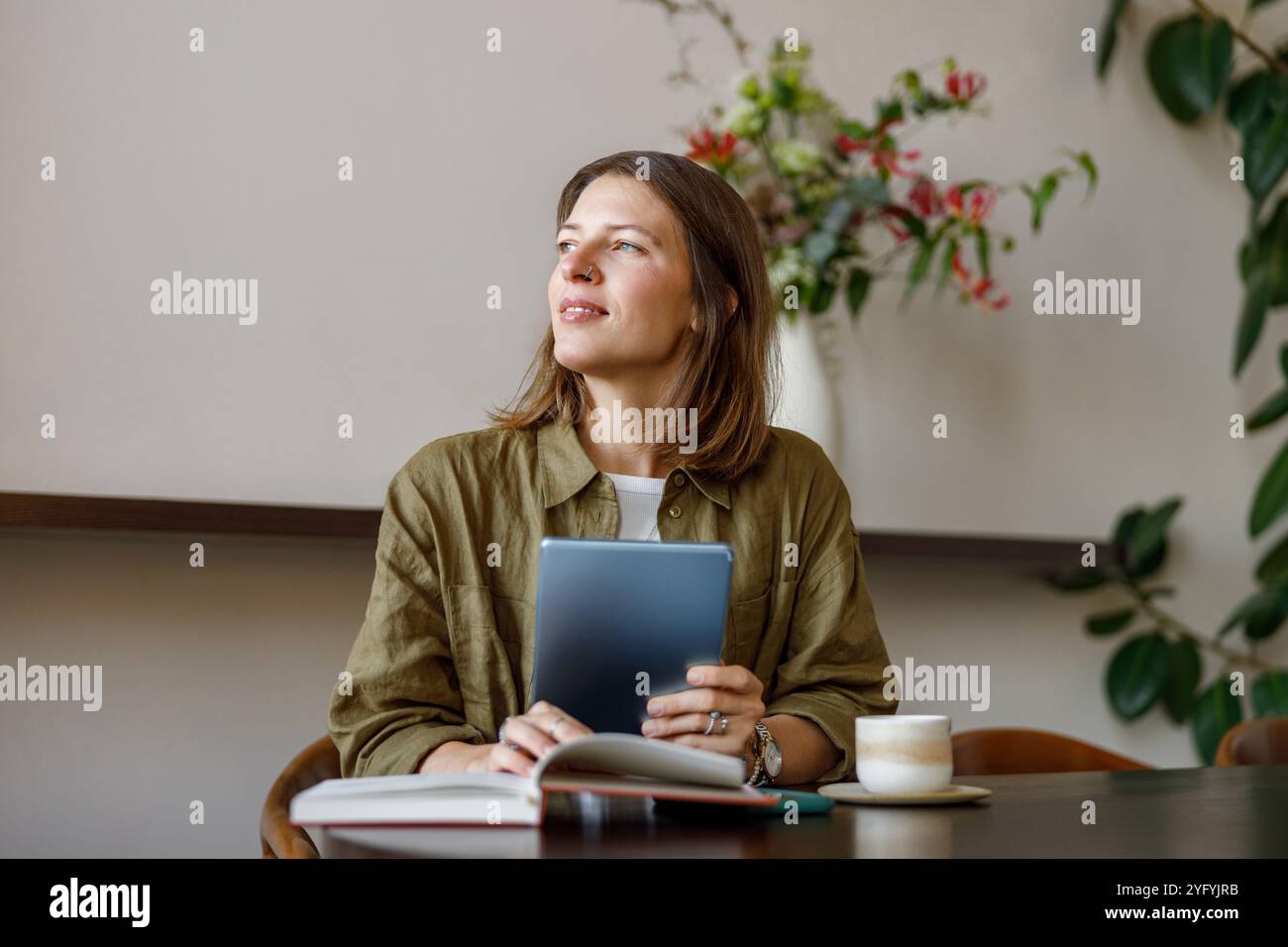 Eine nachdenkliche Frau sitzt in einem gemütlichen Café, während sie ein Tablet hält und sich in Reflexion einlässt Stockfoto Eine nachdenkliche Frau sitzt in einem gemütlichen Café, während sie ein Tablet hält und sich in Reflexion einlässt Stockfoto