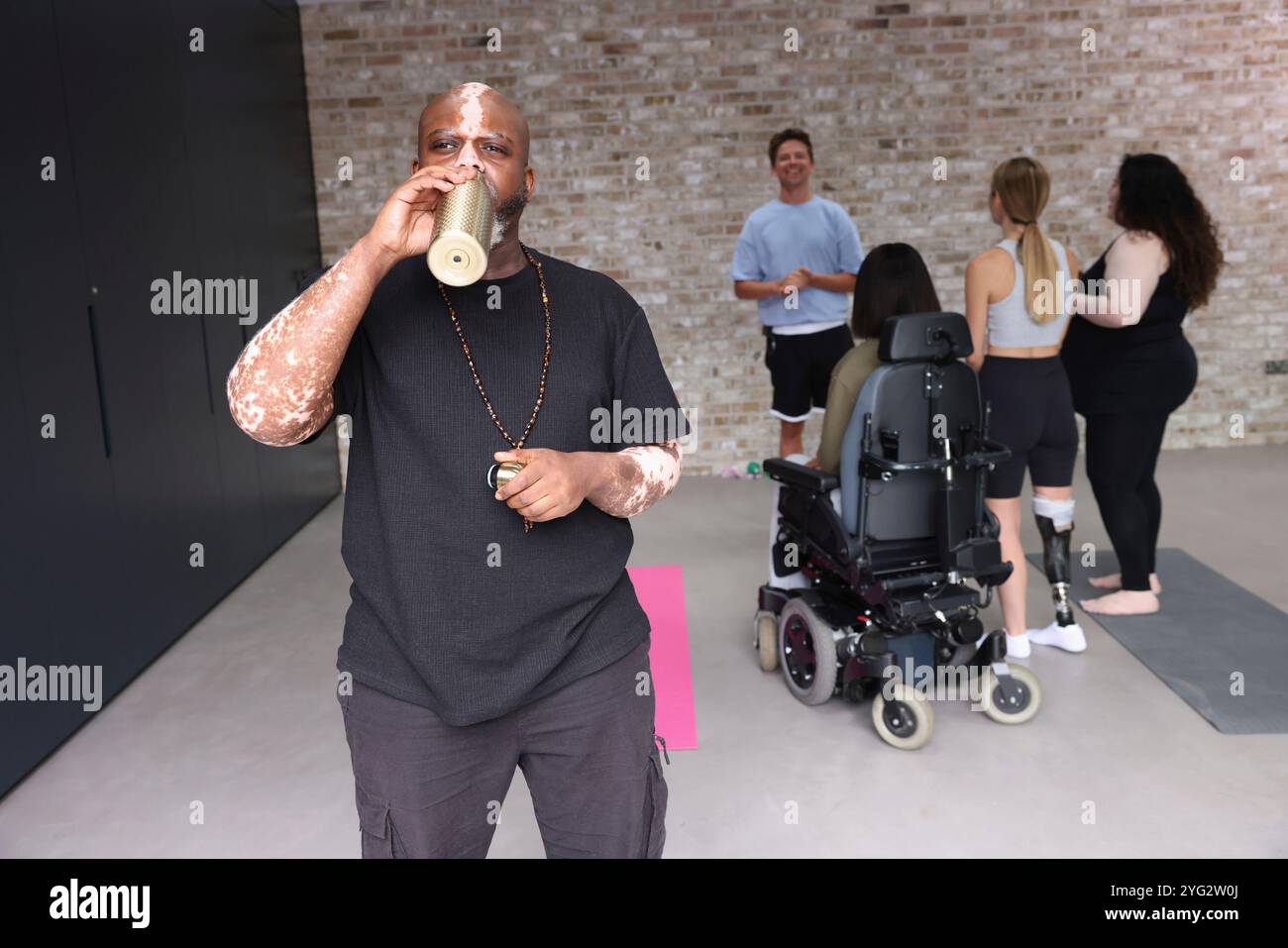 Mann mit Vitiligo, der im Fitnessstudio steht, aus der Wasserflasche trinkt, Leute im Hintergrund Stockfoto
