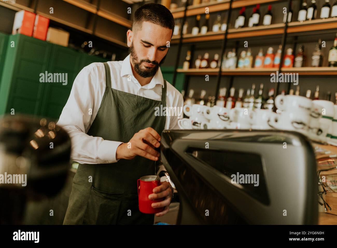 In einem schicken Café konzentriert sich ein erfahrener Barista auf die Herstellung köstlicher Getränke, umgeben von ordentlich angeordneten Gläsern und lebendiger Einrichtung, die eine warme Atmosphäre schaffen Stockfoto