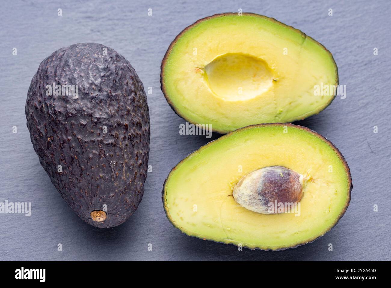 Drei Avocados, eine ganze und zwei Hälften, sind auf einer grauen Schieferoberfläche angeordnet. Die halbierten Avocados zeigen ihr cremiges gelbes Fleisch und ihr großes braunes p Stockfoto
