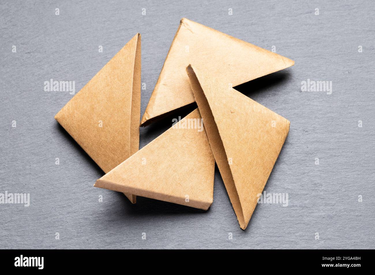 Vier braune Papierdreiecke sind kreisförmig auf einer grauen Oberfläche angeordnet. Die Dreiecke überlappen sich leicht. Stockfoto