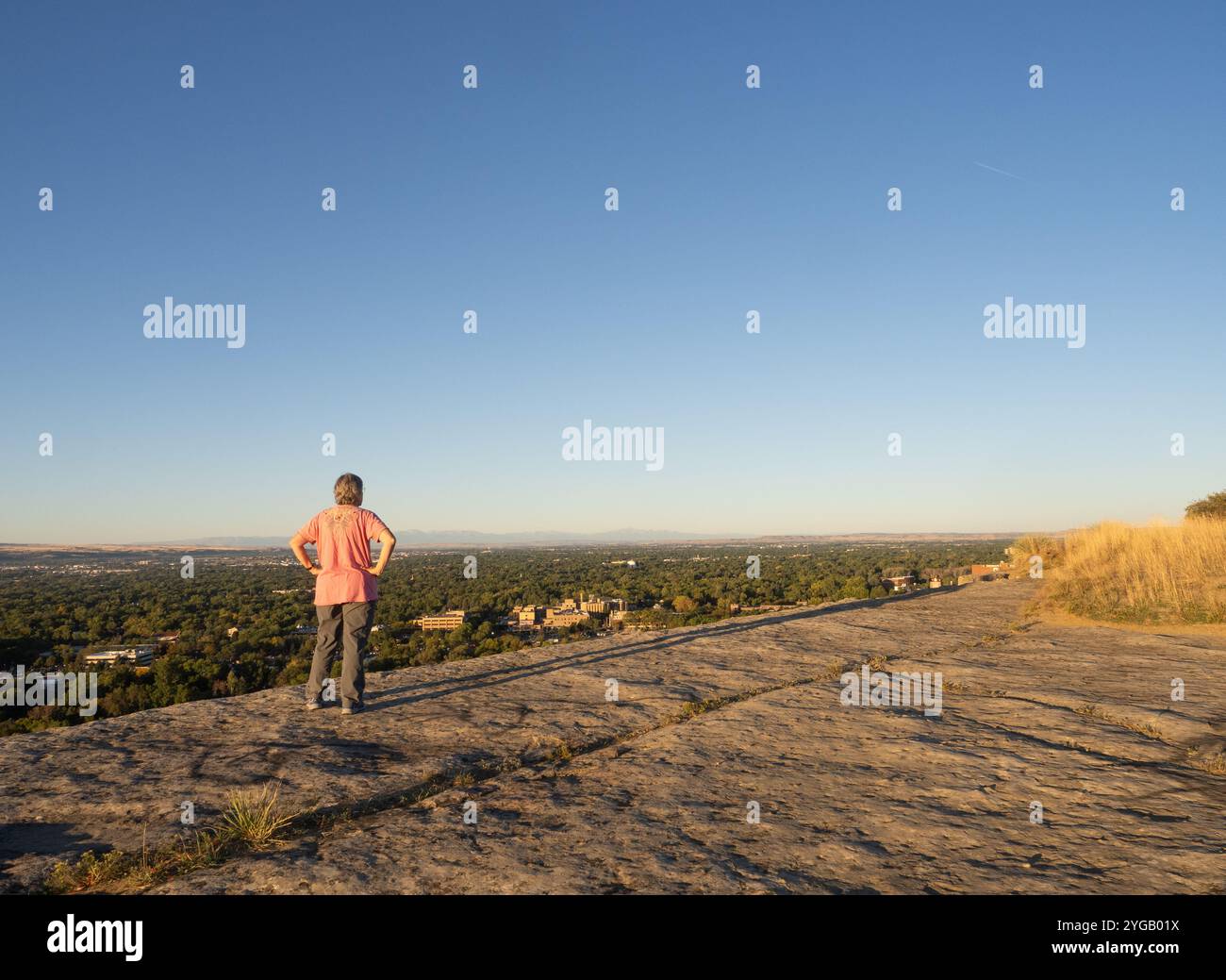 Ältere Frau mit rosa Hemd und grauer Hose auf einem Felskamm mit Blick auf eine Stadt. Fotografiert mit dem Rücken zur Kamera in Billings, Mo Stockfoto