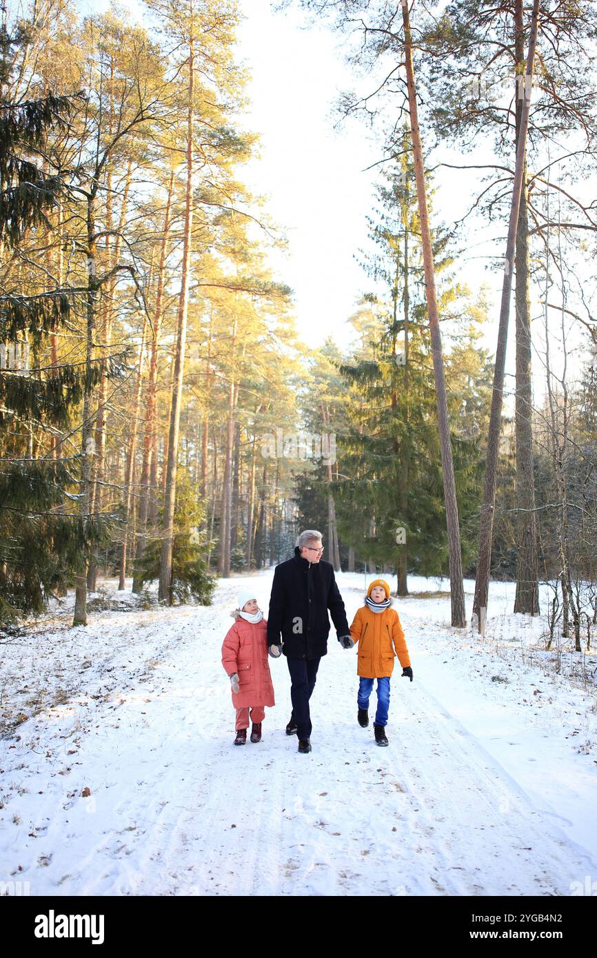 Ein Vater macht einen gemütlichen Spaziergang mit seinen beiden Kindern durch einen wunderschönen verschneiten Wald. Die Szene fängt die Freude der Familie in der ruhigen Winterlandschaft mit schneebedeckten Bäumen ein, die eine magische Kulisse bilden. Stockfoto