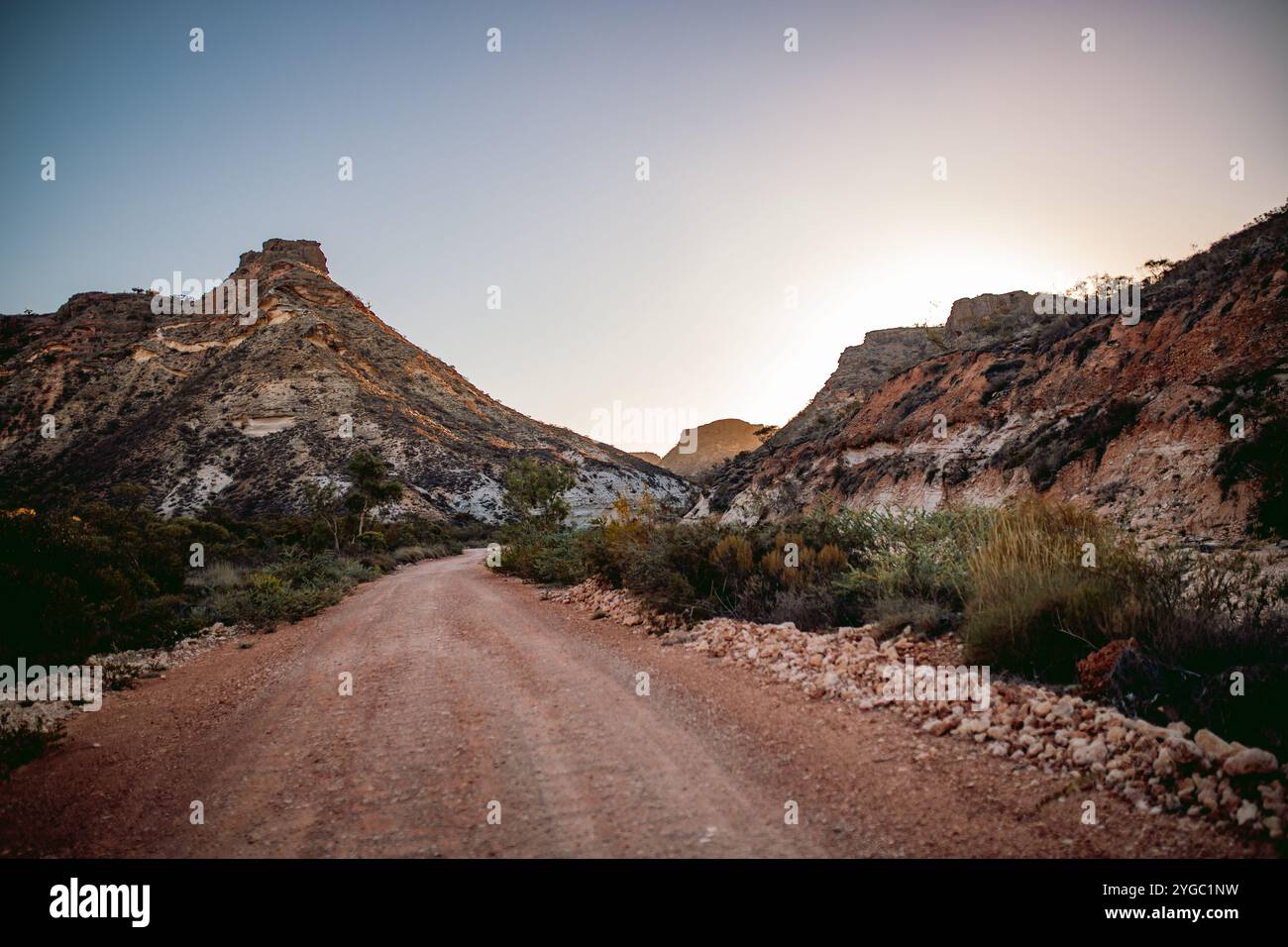 Rote Feldstraße umgeben von australischem Busch, die während der goldenen Stunde zu sonnendurchfluteten Canyonkämmen im Cape Range National Park führt. Goldener Dunst. Stockfoto