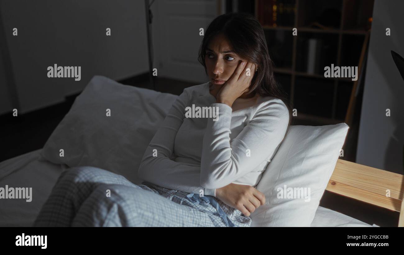 Hispanische brünette Frau sitzt nachdenklich im Bett in einem schwach beleuchteten Schlafzimmer ihres Hauses, trägt bequemen Pyjama und ruht auf einem weißen pi Stockfoto