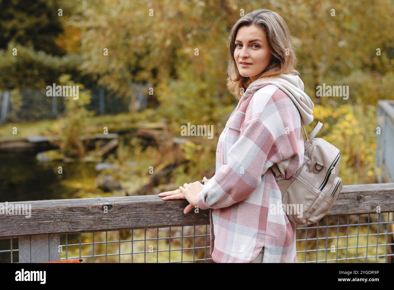 Porträt einer niedlichen jungen Frau in lässiger Kleidung im Herbst, stehend auf der Brücke vor dem Hintergrund eines Herbstparks und Flusses. Hübsche weibliche Spaziergänger in Par Stockfoto