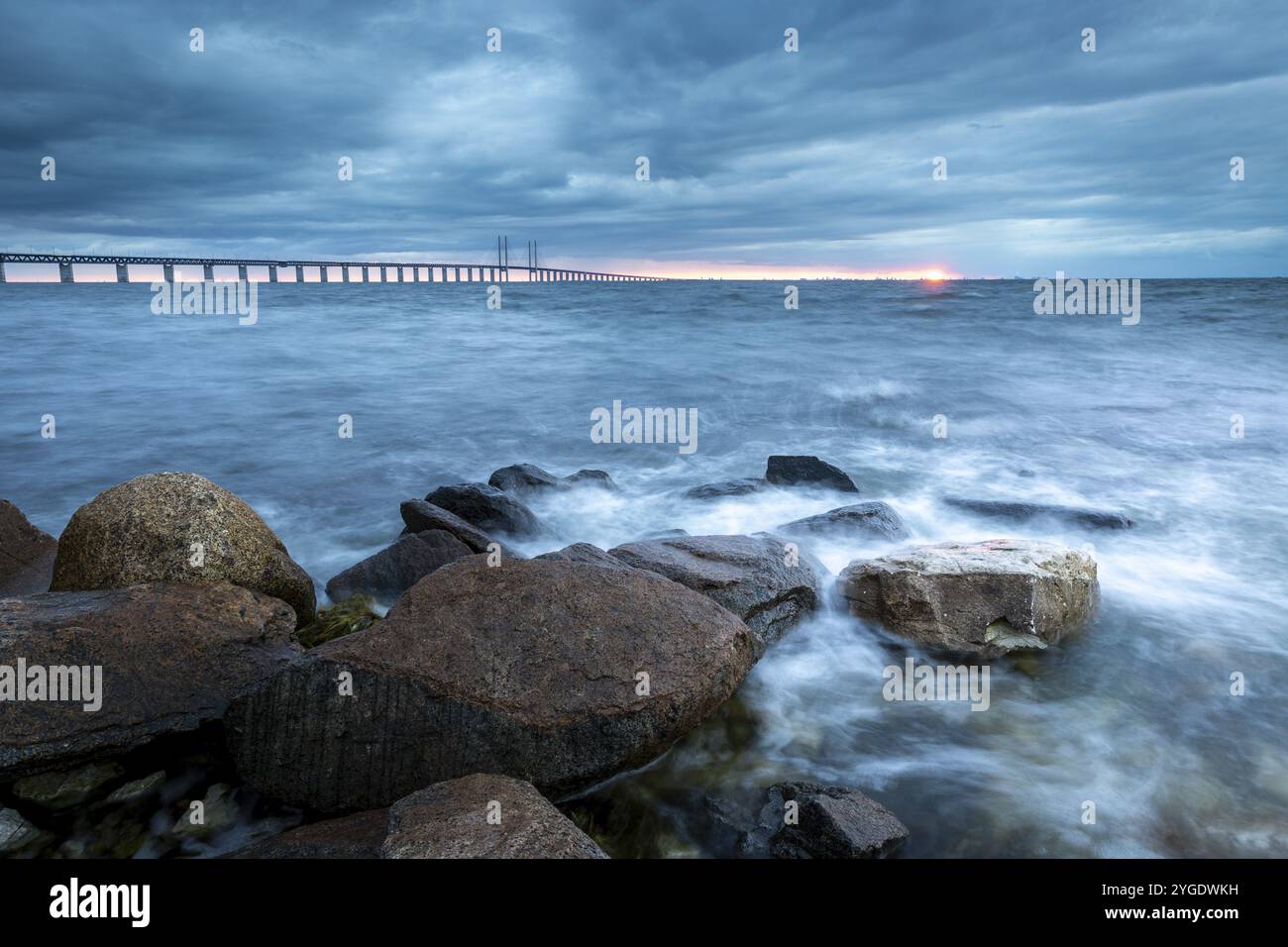 Dramatische Atmosphäre mit Meer, Brücke und Felsen im Vordergrund bei Sonnenuntergang, Oeresund Bridge, Oresundsbroen, die längste Seilbrücke der Welt, Stockfoto