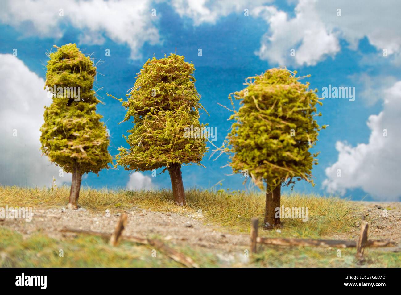 Diorama von drei Bäumen auf einem Hügel mit einem Kiesweg, gebrochenem Holzzaun und teilweise bewölktem Himmel. Stockfoto