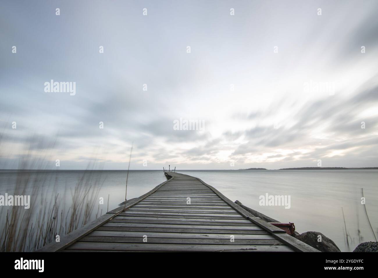 Sonnenaufgang über einem See im Herbst. Ein Holzsteg ragt ins Wasser, während sich Gras am Sandstrand angesiedelt hat. Der See und der Strand am V-See Stockfoto