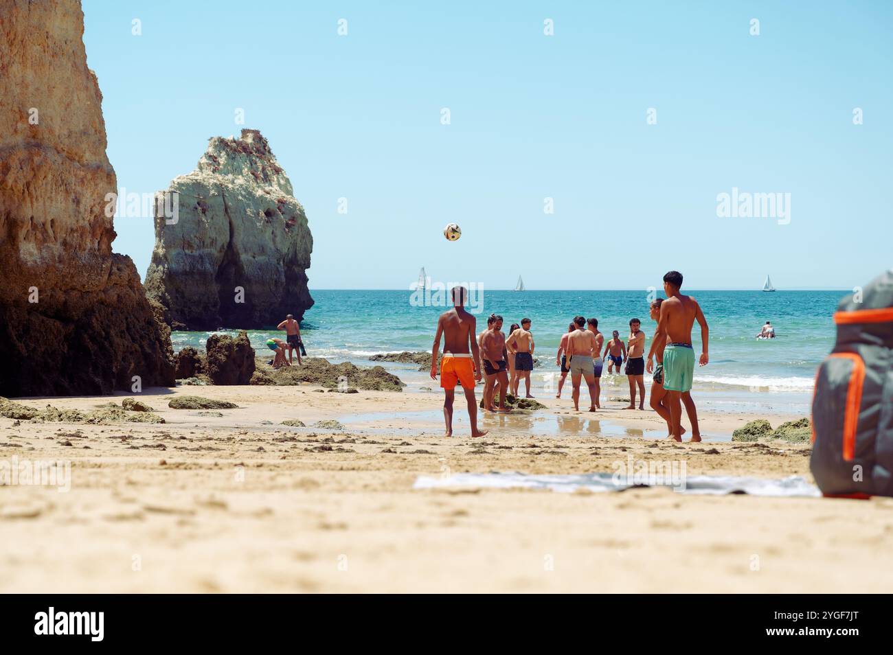 Freunde spielen Strandfußball mit malerischen Klippen und Segelbooten im Hintergrund in praia da rocha Stockfoto