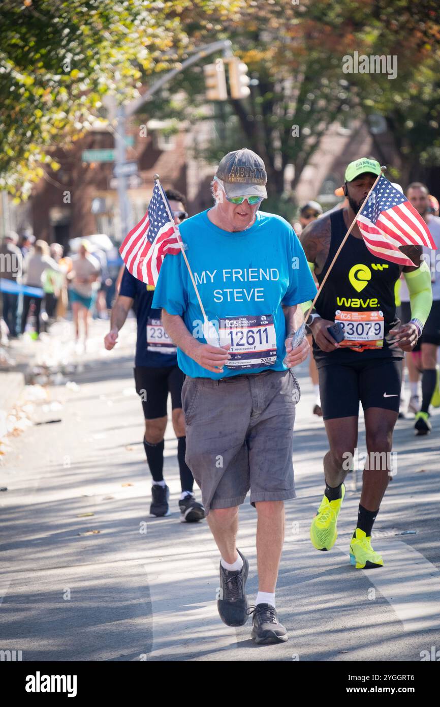 Ein düsterer Läufer beim NY Marathon 2024 mit 2 amerikanischen Flaggen und einem T-Shirt für einen Freund/Verwandten. In Williamsburg, Brooklyn. Stockfoto