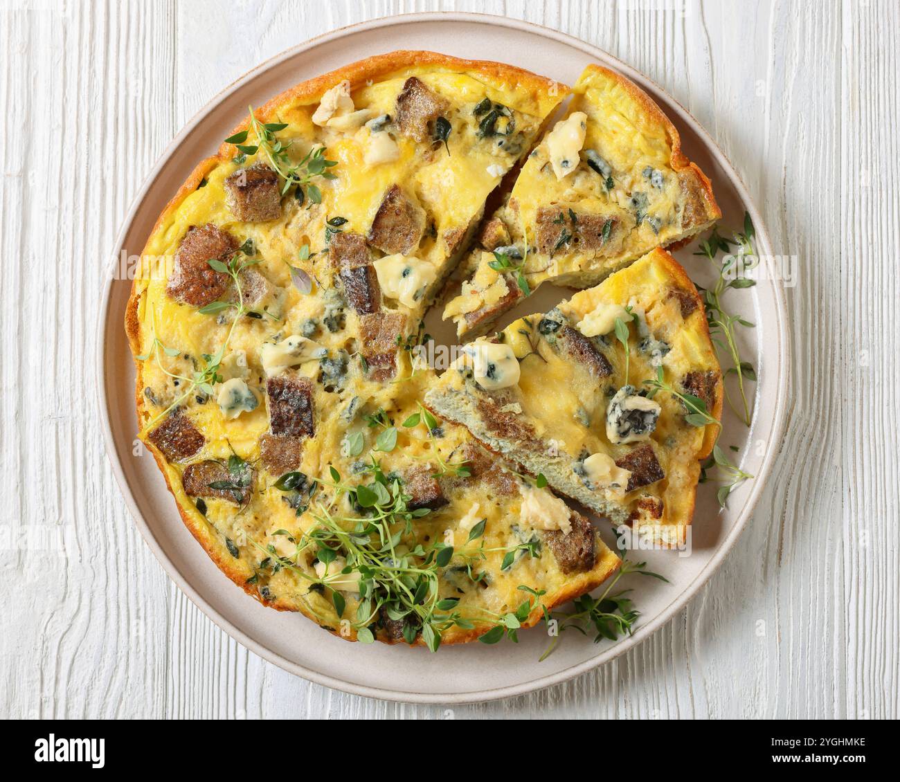 Gebackenes Omelette mit Blauschimmelkäse, Roggencroutons und frischem Thymian auf Teller auf weißem Holztisch, horizontaler Blick von oben, flache Lage, Nahaufnahme Stockfoto