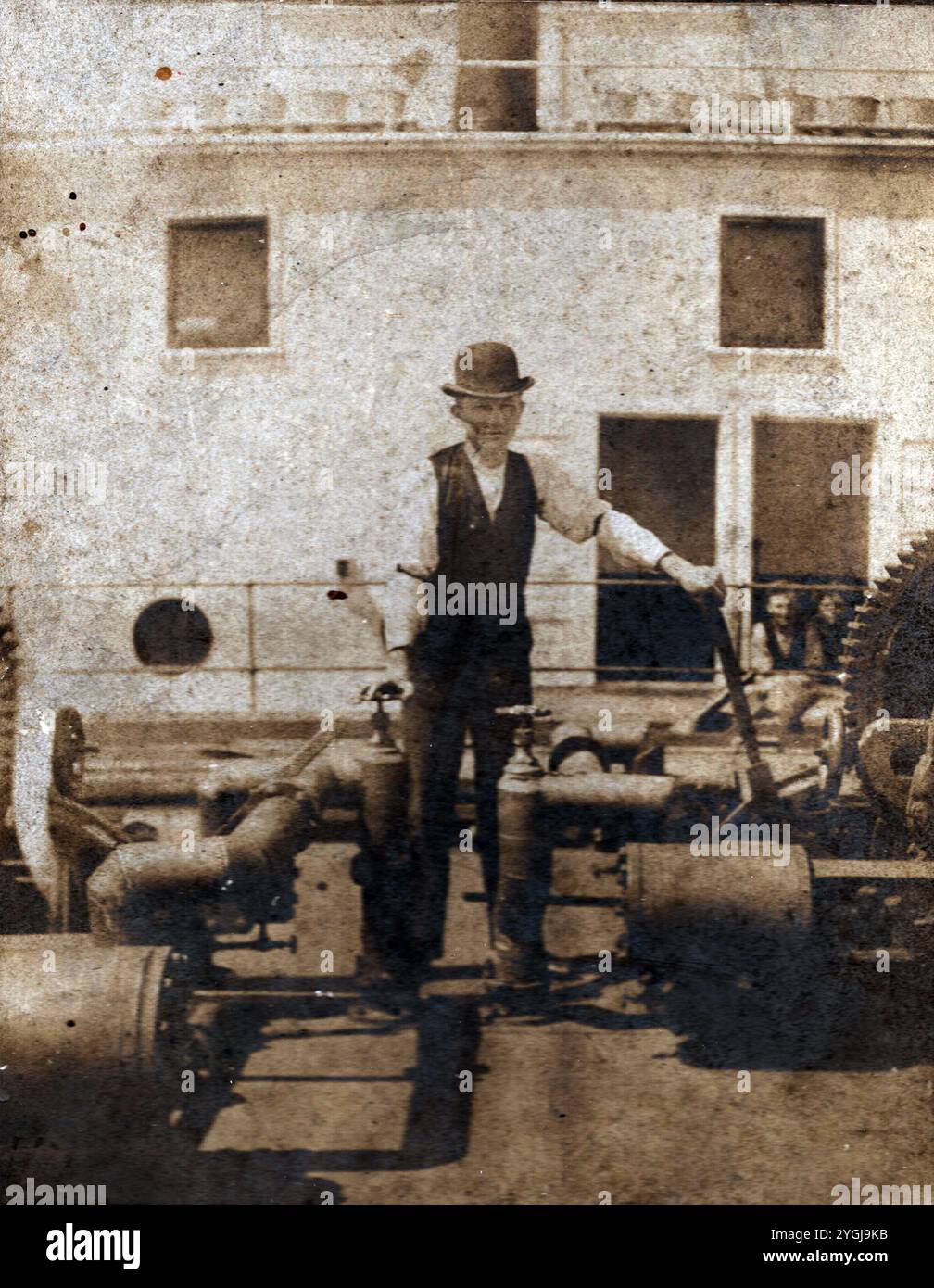 Dies ist ein originelles und echtes Foto eines Mannes in Kleidung aus dem 19. Jahrhundert, einschließlich eines Bowler-Hutes, der neben großen Zahnrädern und Rohren auf dem Deck eines Dampfschiffes steht. Die maritime Umgebung und seine Kleidung deuten auf einen Zeitrahmen der späten 1800er bis frühen 1900er Jahre hin, wahrscheinlich während der Spitzenzeiten dampfbetriebener Schiffe. Das Bild bietet einen seltenen Einblick in die historische Seefahrertechnik und das Leben auf Schiffen und bewahrt ein Stück der Industrie- und maritimen Geschichte dieser Epoche. Stockfoto