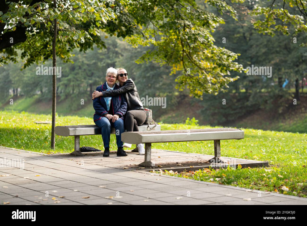 Eine ältere Frau in Blau und ihre Tochter in schwarzem Leder genießen einen Herbsttag auf einer Parkbank. Die zwanglose Szene fängt ihre Familienverbindung ein Stockfoto