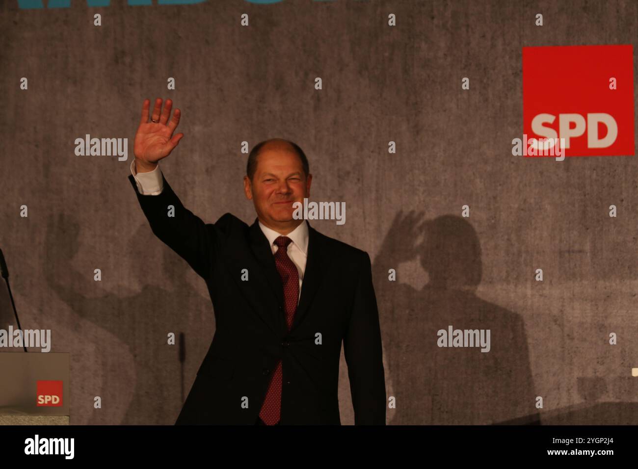 OLAF Scholz steht vor einer grauen Wand und winkt entspannt ins Publikum. Rechts im Bild ist das SPD-Logo. Stockfoto