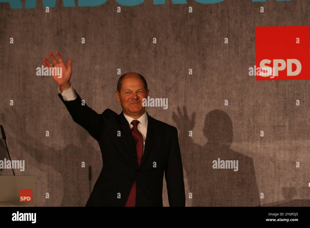 OLAF Scholz steht vor einer grauen Wand und winkt entspannt ins Publikum. Rechts im Bild ist das SPD-Logo. Stockfoto