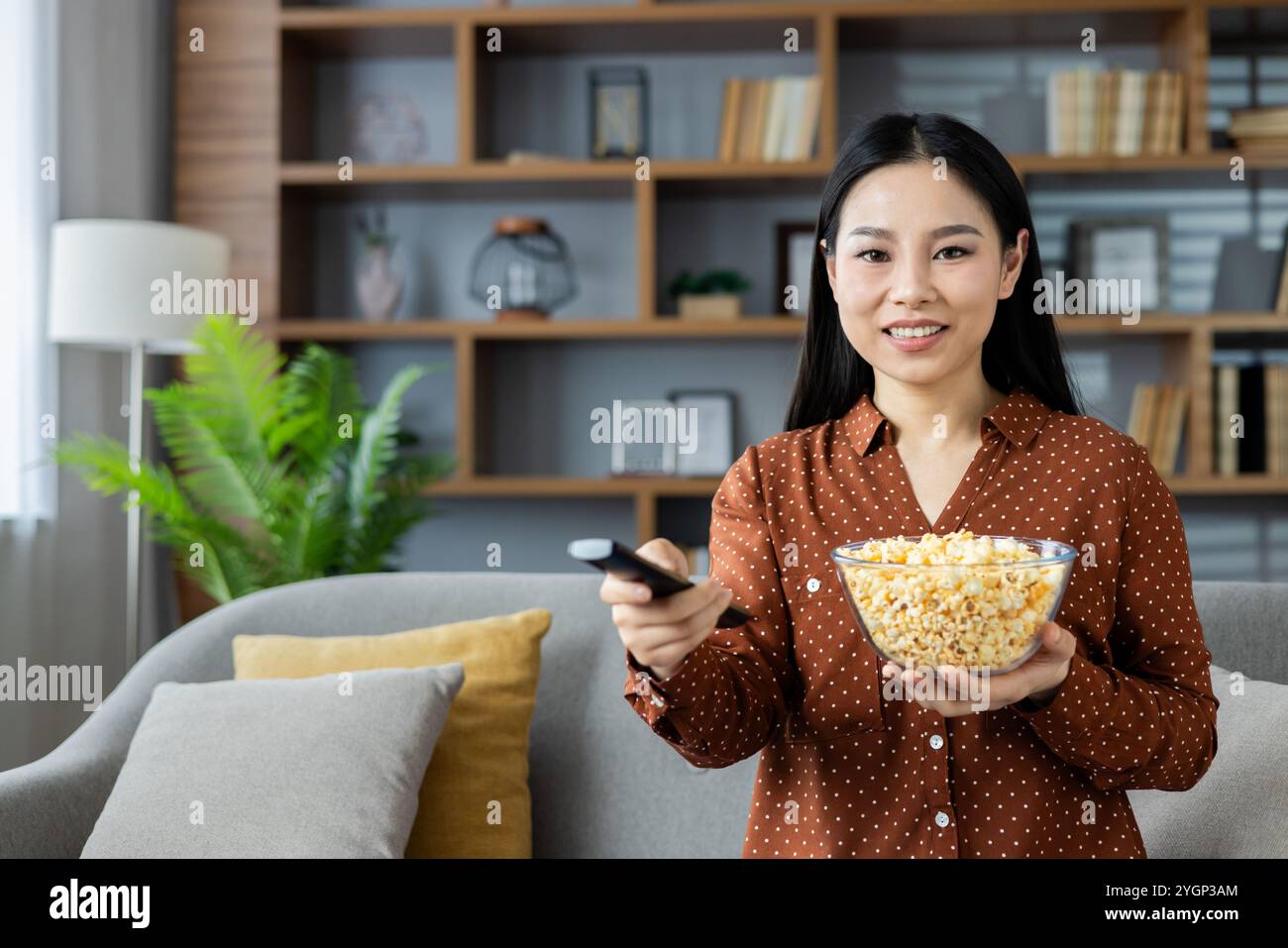 Asiatische Frau, die bequem auf dem Sofa sitzt, Popcorn und Fernbedienung hält. Entspannte Atmosphäre zu Hause mit gemütlichen Kissen, die auf Freizeit schließen lassen. Bereit für den Filmabend, genießt sie drinnen Ruhe. Stockfoto