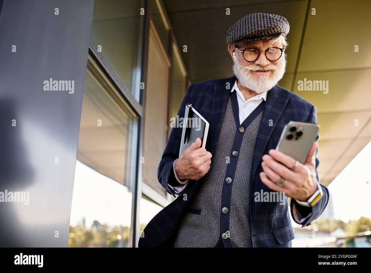 Ein stilvoller Seniorenmann mit Bart überprüft sein Smartphone, während er im Herbst in der Nähe eines modernen Gebäudes steht. Stockfoto