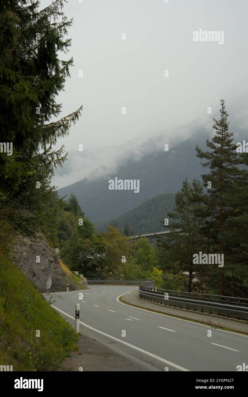 Gewundene Bergstraße durch die Schweizer Alpen an einem nebeligen Tag mit dichtem Grün und bewölktem Himmel Stockfoto