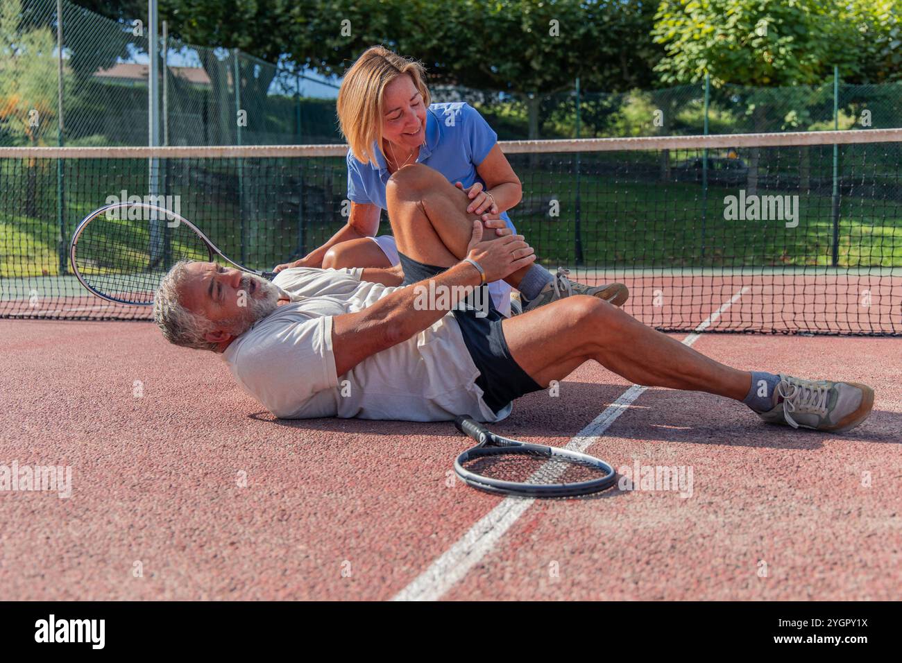 Eine ältere Tennisspielerin kümmert sich um ihre Partnerin, die mit einer Beinverletzung auf dem Tennisplatz liegt und Schmerzen zu haben scheint. Hebt deren Sup hervor Stockfoto