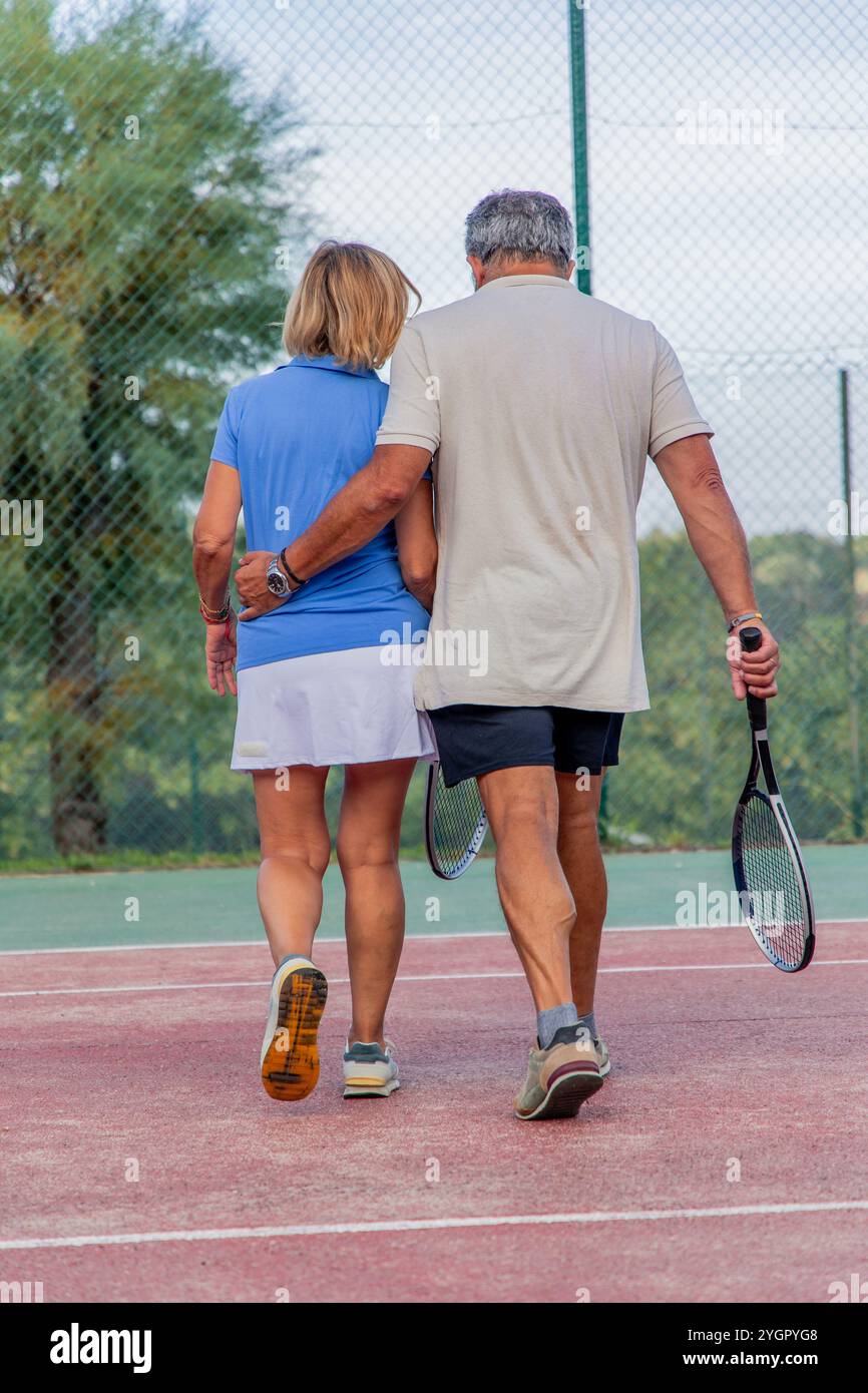 Vertikales Senior Tennis-Paar geht Arm in Arm vom Platz und teilt sich nach einem harten Match einen Moment der Unterstützung. Ihre enge Umarmung spiegelt beide Kompa wider Stockfoto