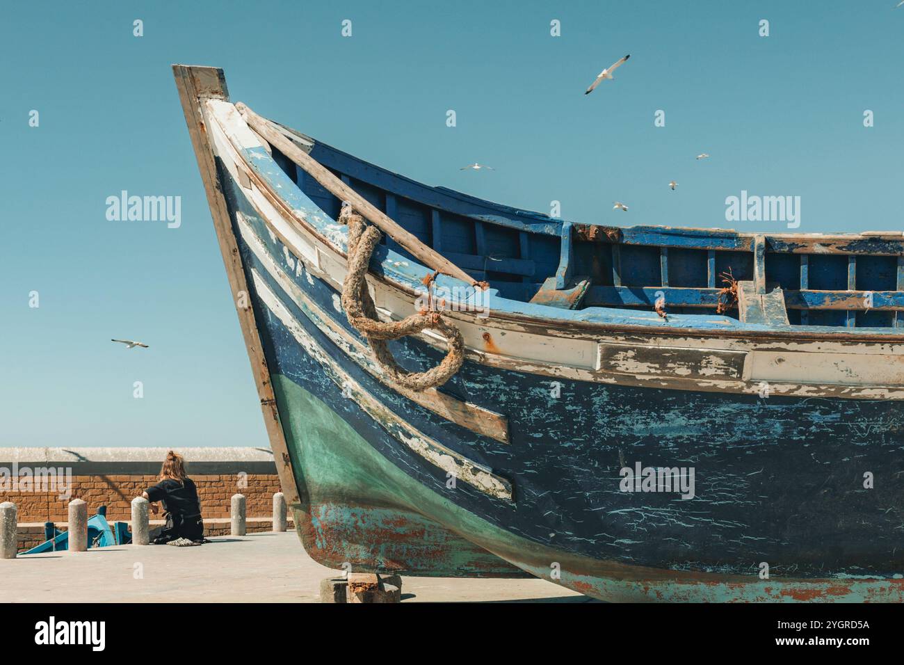 Junge weibliche kaukasische Touristin, die im Hafen von Essaouira sitzt und auf das Meer blickt, die Aussicht mit traditionellen blauen Booten bewundert, Stockfoto