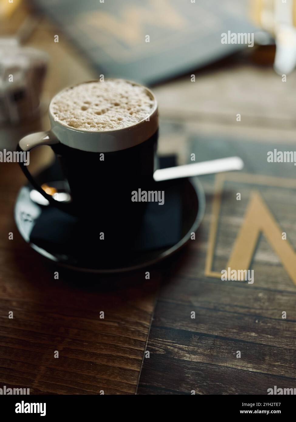 Eine warme Tasse aufschäumenden Kaffee auf einem rustikalen Holztisch mit verschwommenem Hintergrund, der die gemütliche und einladende Atmosphäre unterstreicht. - Smartphone-aufgenommenes Stockfoto