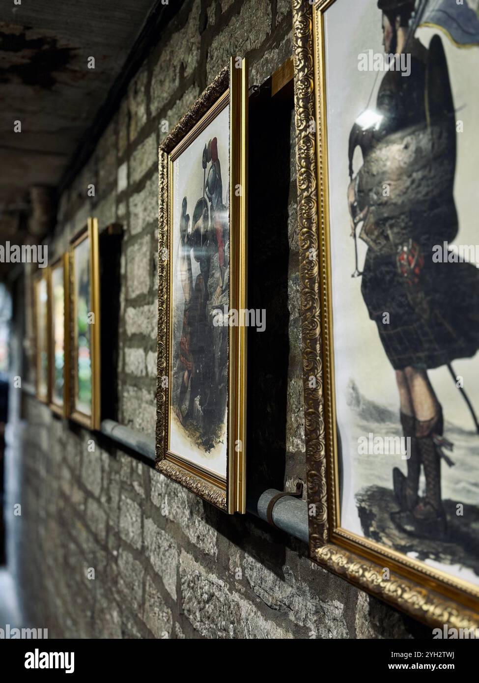 "Eine Reihe historischer Gemälde in kunstvoll verzierten Goldrahmen hing an einer rustikalen Steinmauer in einem schwach beleuchteten Flur." - Smartphone-aufgenommenes Stockfoto