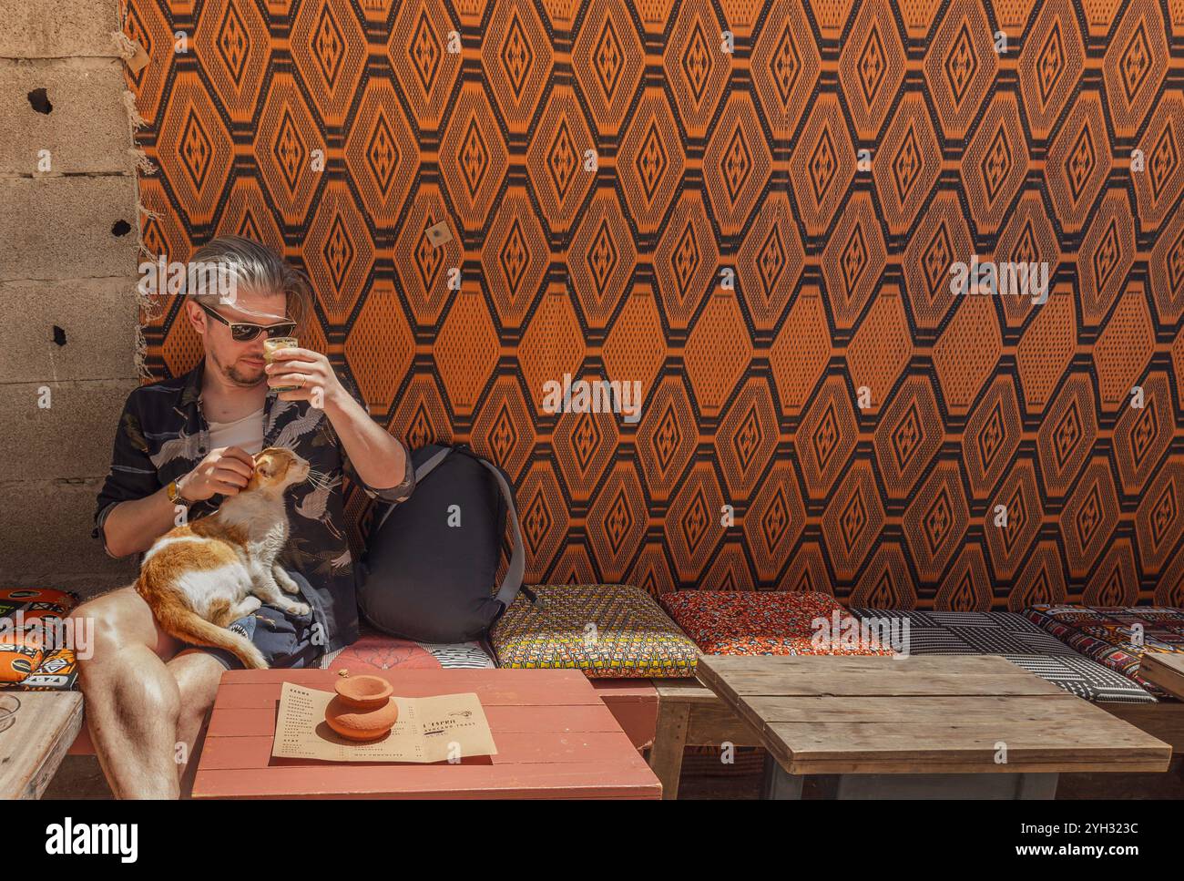 Junger Erwachsener kaukasischer Reisender, der im Essaouira-Café sitzt und einen Kaffee mit einer Katze auf dem Schoß hält, mit lebhaftem orangefarbenem marokkanischem Dekor und Stockfoto